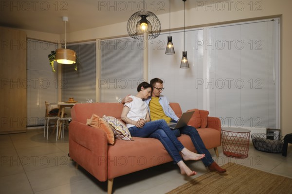 A husband embraces his wife while sitting on a couch in a modern living room. They are watching a
