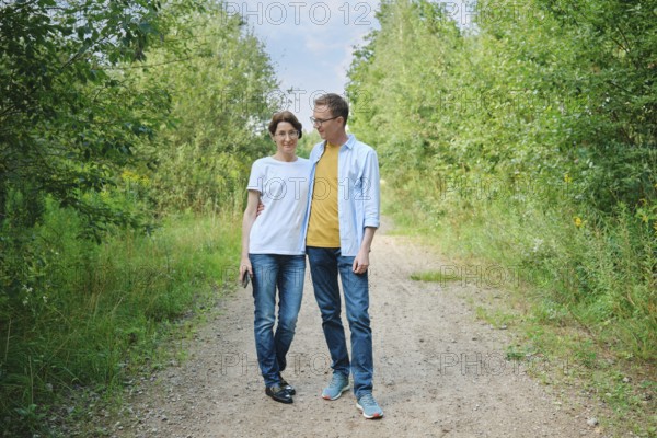A middle-aged couple enjoys a stroll along a gravel path shaded by lush greenery. They smile at
