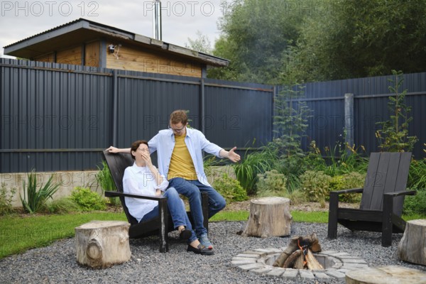 A couple spends a relaxed evening outdoors near a fire pit in their backyard. They share playful