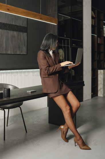 A businesswoman in a brown suit is sitting on a desk while focused on her laptop in a contemporary