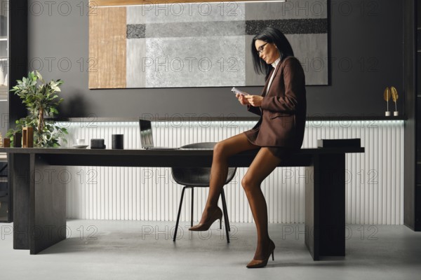 Serious business woman in a brown suit sits on a desk, focused on her smartphone. She checks her