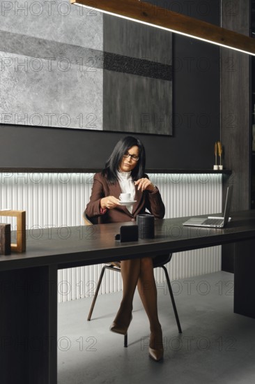 A woman in business attire sits at a sleek desk in a modern office, taking a cup of coffee. The