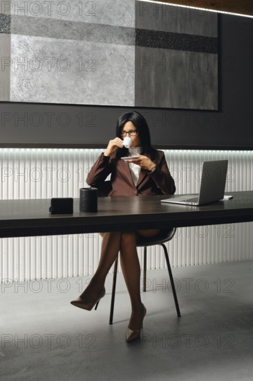 A confident businesswoman is seated at a sleek desk in a contemporary office. She sips coffee while