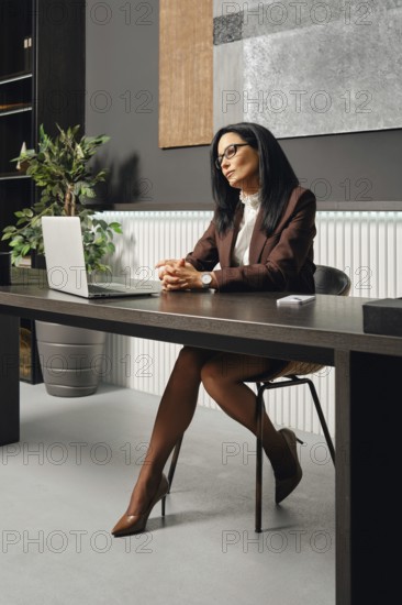 A woman dressed in business attire sits at an office desk in office and looking at the screen of