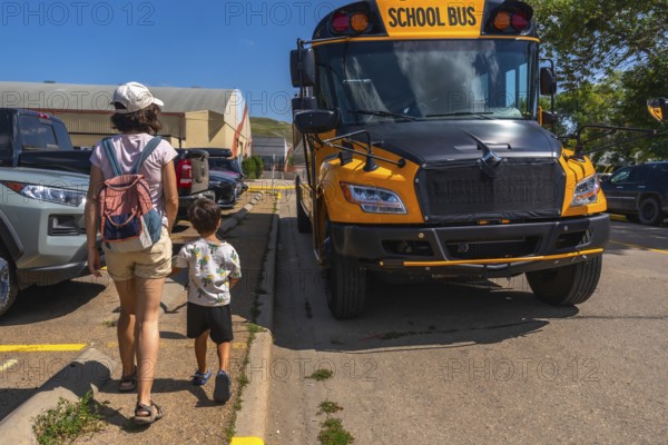 Mother and son walking hand in hand towards a bright yellow school bus in a bustling parking lot,