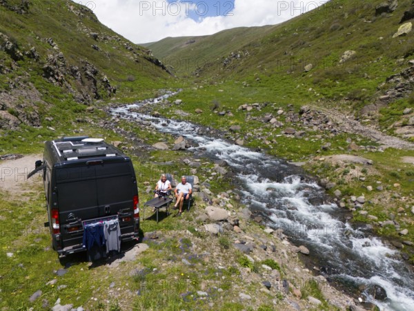 A campervan stands next to a river in the mountains where people sit at a table and enjoy nature,