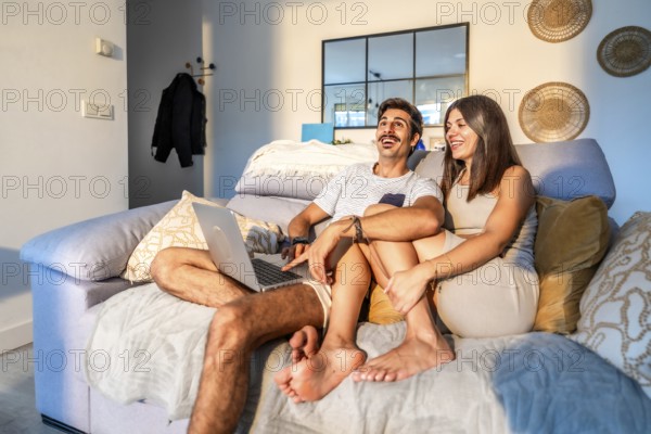 Young couple laughing while watching a streaming movie on their laptop, sitting comfortably on the