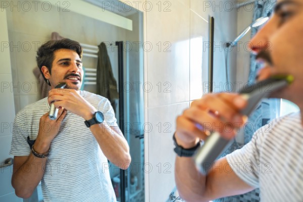 Young man trimming his beard using an electric razor, looking at himself in the bathroom mirror