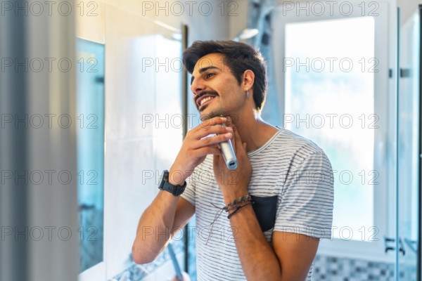 Happy young man trimming his beard in the bathroom mirror using an electric razor, enjoying his