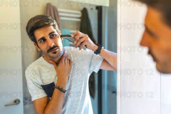 Concentrated young man trimming his beard with electric razor in bathroom mirror, morning routine,