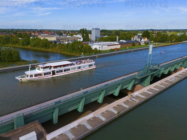 A passenger ship sails on a canal over a large bridge, surrounded by city buildings and nature,
