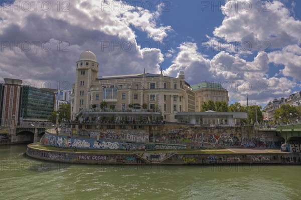 A historic building by the river with graffiti, under a clear sky, surrounded by urban surroundings