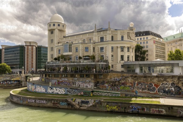 A historic building on the riverbank with graffiti on the wall and a cloudy sky Urania Ringstraße
