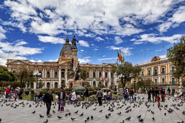 Main square full of people and pigeons in front of a large building with a statue under a blue sky,