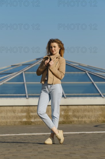 A young woman stands outdoors, wearing a light jacket and jeans. She holds her jacket gently while