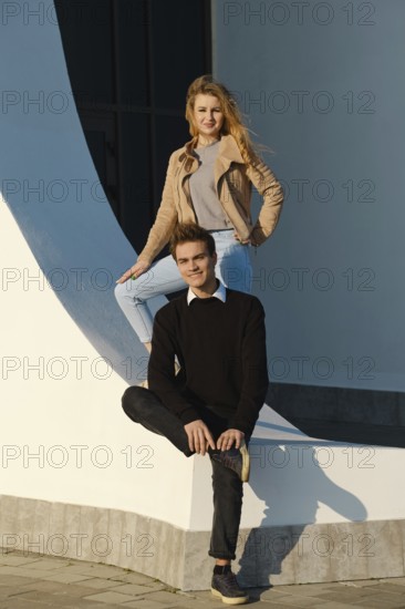 A young man sits on a low wall with his leg bent while a woman stands above him, both smiling. The