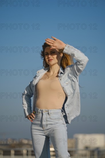 A woman stands outdoors on a sunny day, shielding her eyes from the sunlight. She wears a light