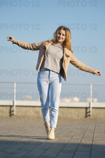 A young woman stands on one foot with arms outstretched, smiling under a clear blue sky. She wears