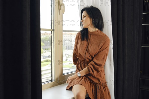 A woman in a brown dress relaxes by a large window, gazing thoughtfully outside. Sunlight filters