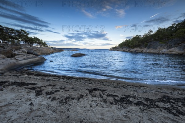 Small bay with sand and rocks, surrounded by pine trees under a partly cloudy sky, Skjeberg,