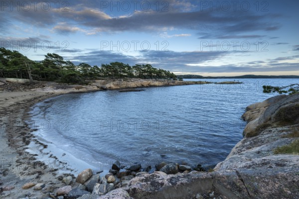 Small bay with sandy beach, surrounded by rocks and pine trees under a partly cloudy sky, Skjeberg,