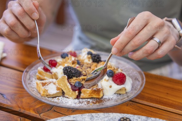 Hands holding spoons over waffles with cream and berries on a table