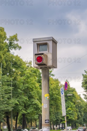 Traffic surveillance camera on a pole, surrounded by trees and road signs under a cloudy sky,