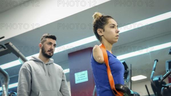 A young female athlete with limb loss trains in a fitness gym with the support of her coach,
