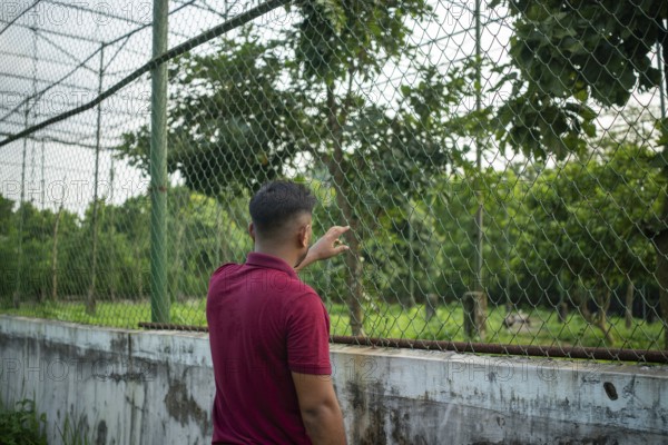 A man stands by a chain-link fence surrounded by lush greenery, appearing contemplative-Dhaka,