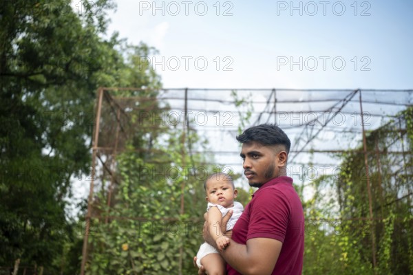 A man holds a baby outdoors by a metal structure with greenery in a relaxed setting-Dhaka,