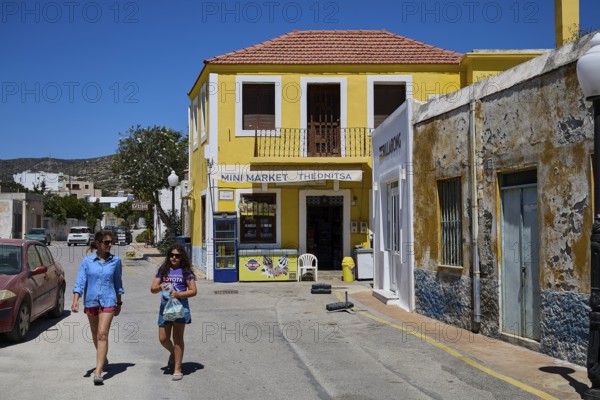 Two woman in summer clothes walking along a yellow building, Kattavia village, southern tip of