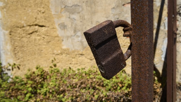 Rusted lock on a metal post in front of a weathered wall with plants in the background, Old silk