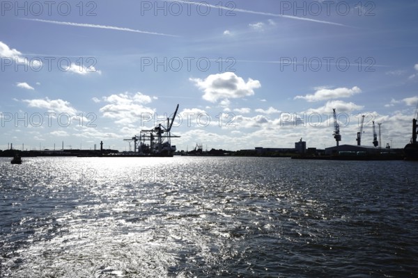 An industrial harbour with cranes and reflecting sunlight on the water under a blue sky. The view