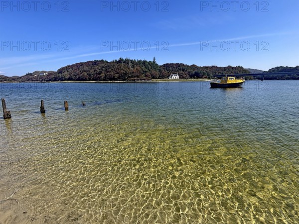 Yellow boat and old wooden piles in a loch with wooded island in the background under blue sky,