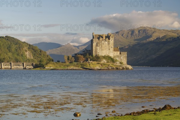 A picturesque castle on an island in the water, surrounded by mountains and a slightly cloudy sky,