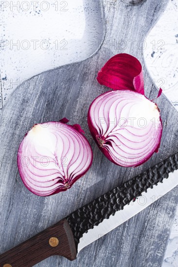 A vibrant red onion is cut in half, on a rustic wooden chopping board beside a sharp knife, ready