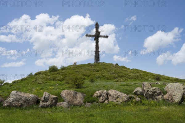 Metal cross on a green hill under a blue sky, surrounded by large stones, Monument to the Holy