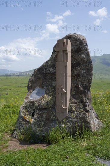 Stone with cross relief in front of a vast green landscape and blue sky, at the Monument of the