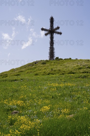 Large metal cross on a hill with a green meadow and yellow flowers under a blue sky, Monument to