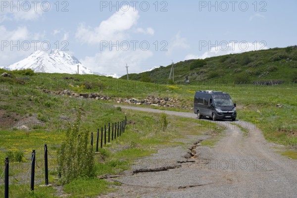 A car drives on a rural road in front of a snow-covered mountain under a blue sky, camper drives
