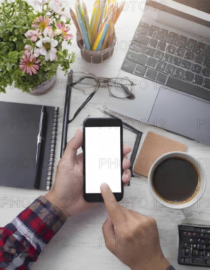 A person is using a cell phone on a desk with a laptop and a cup of coffee. The desk is cluttered