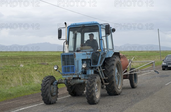 A blue tractor is driven by a man on a country road with fields and cloudy sky, Belarus MTZ-82