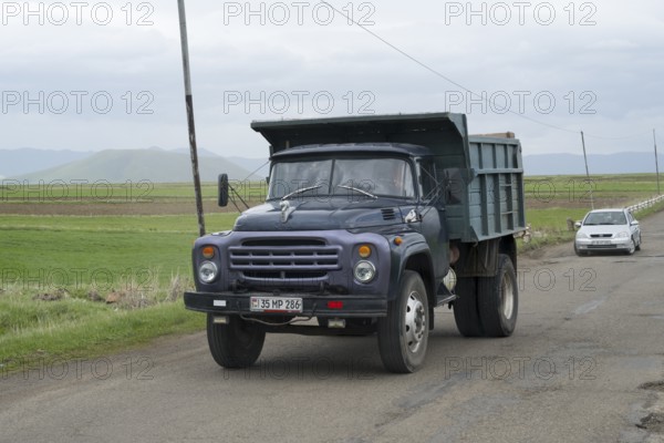 A blue lorry with a trailer drives on a country road surrounded by green fields and mountains,