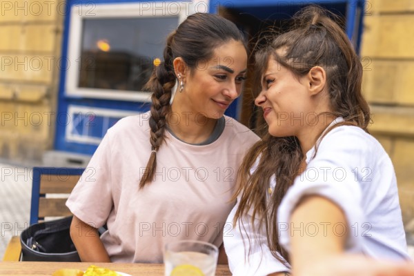 Two women are sitting close together at a table outside a cafe, smiling and enjoying each other's