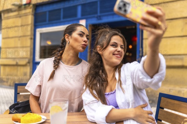 Two young women are taking a selfie with a smartphone while sitting at an outdoor table, enjoying