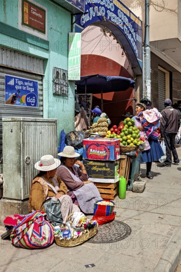 Women in traditional dress sell fruit at a busy market stall in a Bolivian town, selling goods in