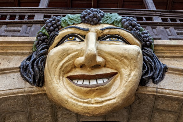 Stone mask with smiling face on an architectural structure, surrounded by green and brown tones,