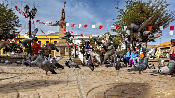 Pigeons fly over a busy square with a monument and garlands under a blue sky, The city pigeons of
