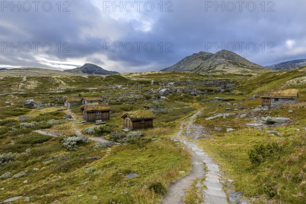Wooden huts in an open meadow in a remote mountain landscape, with winding paths and clouds in the