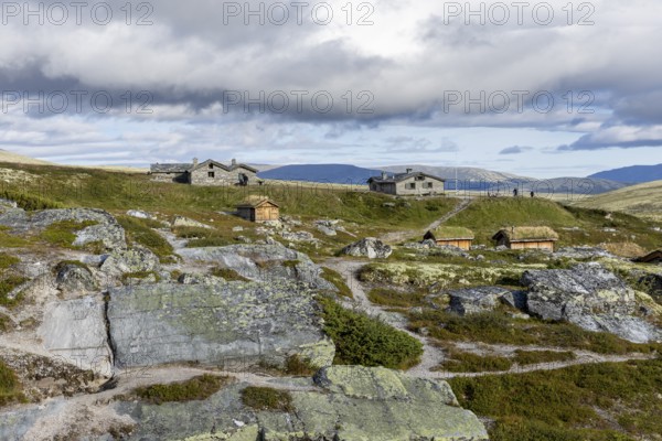 Various wooden cabins on a rocky plain, surrounded by grass and mountains under a cloudy